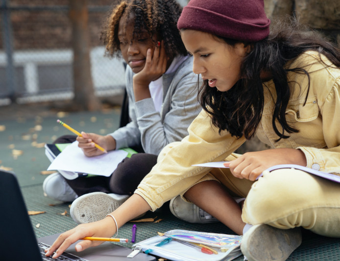 Two students are sitting on the ground with pens and notebooks working at a laptop.